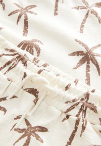 Bijela tkanina s uzorkom palminog drveta u smeđoj boji. Materijal ima teksturiranu, rebrastu završnu obradu s elastičnim pojasom i podesivim vezicama.