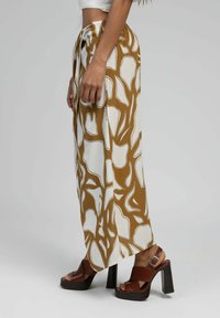 Pantalons larges dans un motif abstrait marron et blanc, en tissu léger. Présentent une taille à cordon de serrage et des fentes sur les côtés. Associés à des sandales à talons marron.