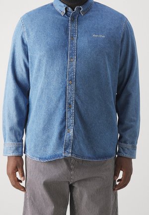 Mężczyzna noszący niebieską jeansową koszulę zapinaną na guziki z logo "Marc O'Polo" na piersi oraz czarno-białe pionowo paskowane spodnie.