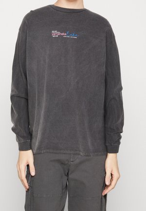 T-shirt en coton à manches longues, gris foncé, présentant un imprimé graphique subtil en texte bleu et rouge sur le haut à gauche de la poitrine. Encolure ronde, coupe décontractée.