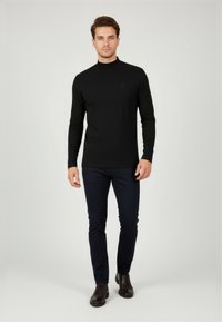 Schwarzes Langarmhemd mit hohem Kragen, figurbetontem Schnitt, kombiniert mit dunkelblauen Hosen und braunen Schnürschuhen. Schlichte Logodetail auf dem Hemd.