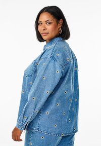 Giubbotto di jeans di colore azzurro chiaro con dettagli circolari ricamati. Presenta un colletto, tasca anteriore e polsini con bottoni. Vestibilità rilassata.
