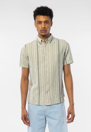 Mann med kortermet skjorte med vertikale grønne, hvite og grå striper, og lyseblå jeans mot en ensfarget bakgrunn.