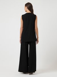 Mulher vestindo uma túnica preta sem mangas e calças largas pretas, de pé contra um fundo claro e liso, de costas.