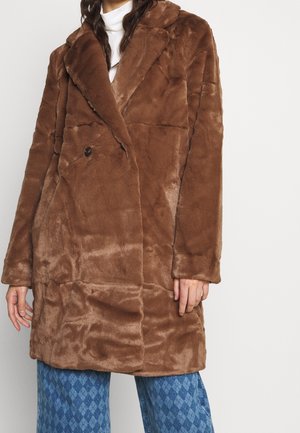 Žena v hnědém plyšovém dvouřadém kožichu z umělé kožešiny přes bílý top a modré kalhoty se vzorem, stojící před jednoduchým pozadím.