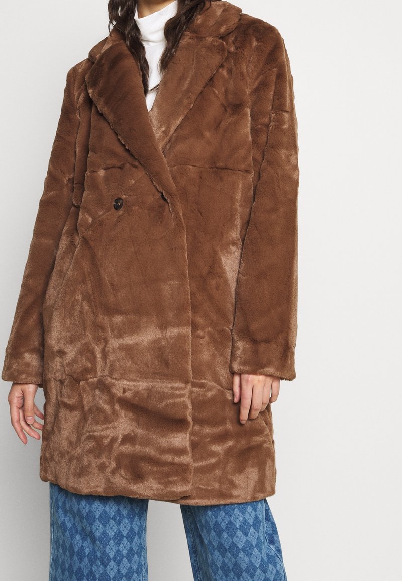 Mujer con un abrigo de piel sintética marrón, de doble botonadura y felpa, sobre una camiseta blanca y pantalones azules con estampado, posando frente a un fondo liso.