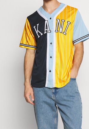 Kurzärmeliges Hemd mit Knopfleiste mit schwarzen, hellblauen und gelben vertikalen Streifen und dem Schriftzug „KANI“, getragen zu hellblauen Jeans.
