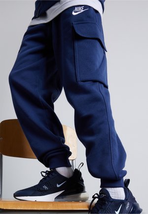 Persona che indossa pantaloni cargo da jogging Nike blu navy e scarpe Nike Air Max 270 nere abbinate, in piedi su una sedia di legno.