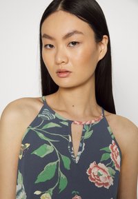 Top halter floral em azul-marinho com decote em gota. Apresenta uma estampa botânica de rosas cor-de-rosa e folhas verdes, tecido texturizado e acabamento contrastante.