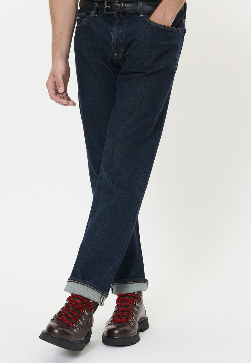 Mand iført mørkeblå, opsmøgede jeans og brune læder vandrestøvler med røde snørebånd, står med krydsede ben mod en ensfarvet baggrund.