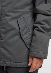 Siva, izolirana jakna z stransko žepom, vidnimi šivi in teksturirano tkanino. Vključuje vrvico na robu.