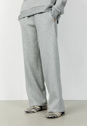 Oseba, oblečena v široke sive trenirke z rokami v žepih in obuta v ravne čevlje z zebra potiskom, stoji na gladki površini.