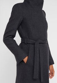 Manteau en laine gris foncé avec ceinture, col montant, poches latérales et manches longues, porté par une personne avec une main dans une poche.