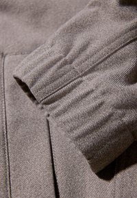 Zbliżenie beżowego rękawa z elastycznym mankietem i widocznymi ściegami na teksturowanym tle odzieży.