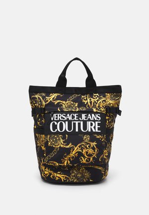 Czarna torba typu tote z złotymi barokowymi wzorami i białym logo "Versace Jeans Couture" na przedniej kieszeni, z dwoma czarnymi uchwytami.