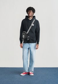 Μαύρο φούτερ με κουκούλα και λογότυπο, ανοιχτόχρωμα μπλε τζιν και κόκκινα αθλητικά παπούτσια. Μαύρη τσάντα χιαστί με λευκές λεπτομέρειες, σε απλό φόντο.