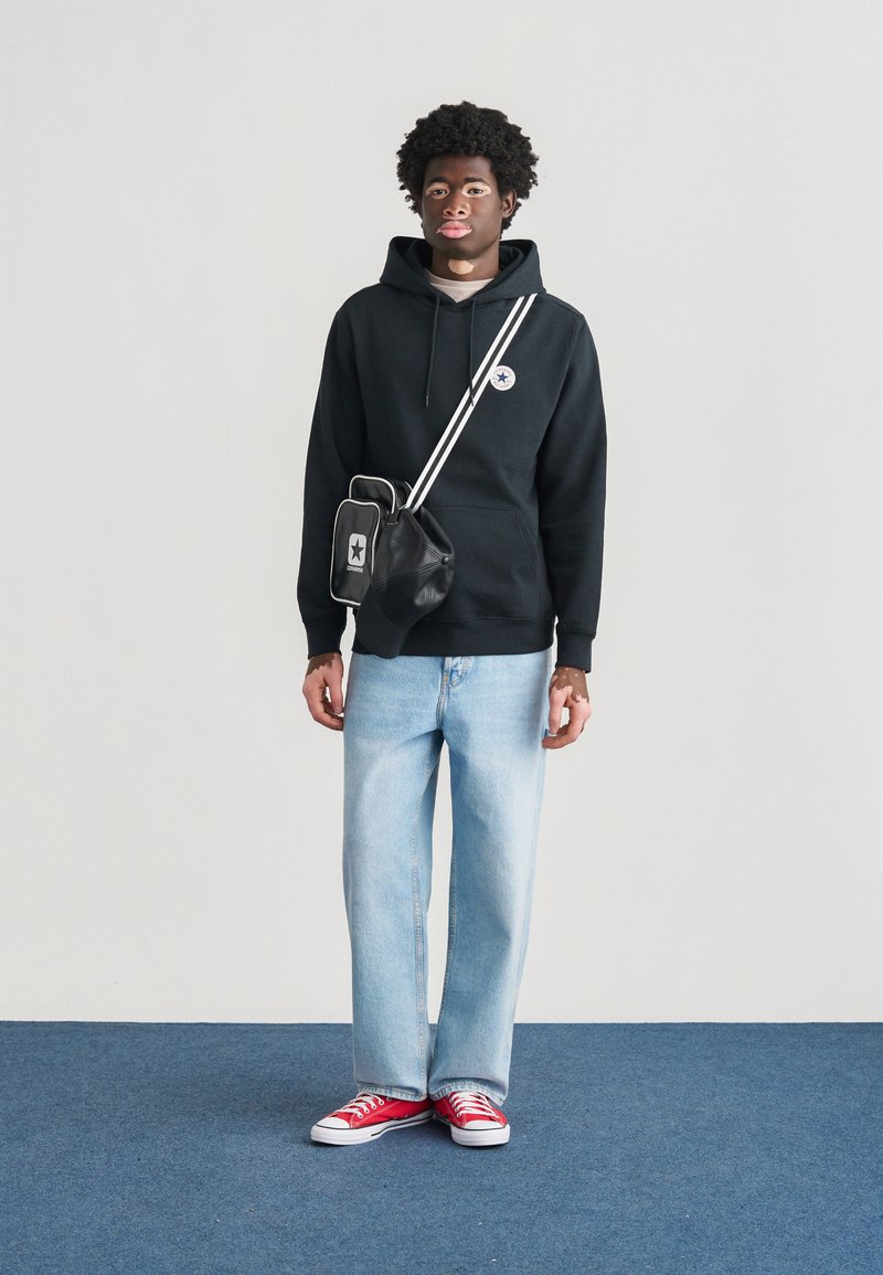 Sort hættetrøje med logo, lyseblå jeans og røde sneakers. Sort crossbody-taske med hvide detaljer, sat op mod en ensfarvet baggrund.