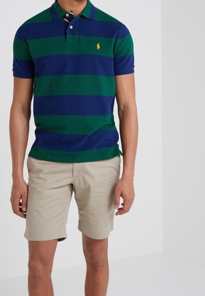 Homme portant un polo rayé vert et bleu marine et un short beige, debout devant un fond blanc uni.