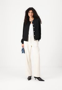 Black textured jacket with silver buttons, white lace top, and cream striped wide-leg trousers. Holding a small blue clutch bag. Black pointed heels.