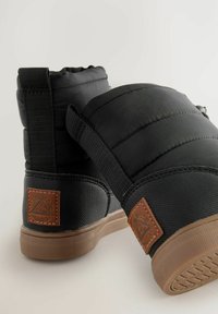 Schwarze gepolsterte Knöchelstiefel mit elastischen Seitenelementen, strukturiertem Gummisohle und einem braunen Lederlogo-Patch an der Ferse.