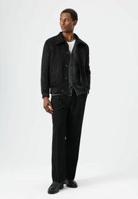 Zwart jack met gestructureerde stof en oversized kraag, gelaagd over een grijze cardigan en wit overhemd, gecombineerd met zwarte broek en schoenen.