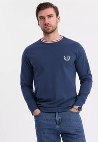 Navy long-sleeve shirt with red and white striped collar, emblem featuring "EX" on left chest, smooth fabric, worn with blue jeans.