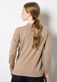 Beige sweater met een ronde halslijn, lange mouwen met kleine hartjespatronen in het wit en bordeauxrood, en ribgebreide manchetten en zoom.