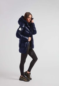 Marineblå dunjakke med hætte, blank finish, quiltet design og en geometrisk lap på ærmet. Kombineret med sorte leggings og sportssko.