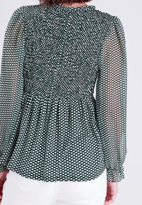 Blouse à motifs verts et blancs avec dos froncé, manches longues transparentes et design plissé, dotée d'un col rond et d'une taille cintrée.