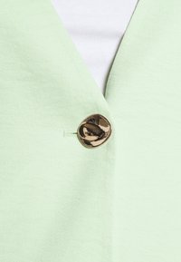 Jasnozielona marynarka o fakturowanym wykończeniu, z złotym guzikiem i dekoltem w kształcie litery V. Materiał jest gładki i miękki.