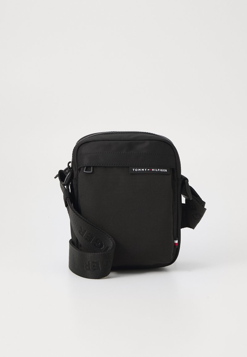 Mala crossbody preta feita de tecido durável, com fecho em zip no topo, patch com logótipo e uma alça ajustável com detalhe da marca.