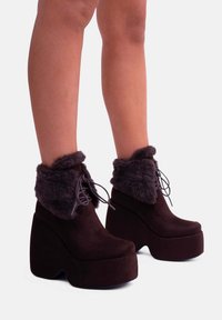 Stivaletti alla caviglia con piattaforma marrone e risvolti in pelliccia sintetica, con chiusura a lacci e finitura liscia e testurizzata. Tacchi a zeppa elevati per un'altezza extra.