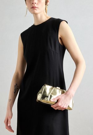 Femme portant une robe noire sans manches et tenant une petite pochette dorée brillante sur fond gris.