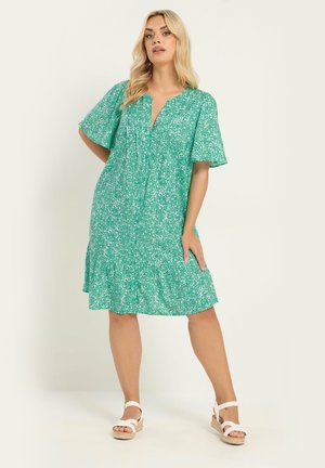 Lyshåret kvinde iført en løs, grøn knælængde kjole med blomsterprint og hvide kilehælsandaler, stående med den ene hånd på hoften.