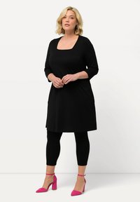 Schwarzes knielanges Kleid mit quadratischem Ausschnitt, dreiviertelärmeln und seitlichen Taschen, kombiniert mit schwarzen Leggings und rosa Absatzschuhen.