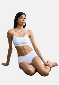 Set di bralette bianca e slip hipster realizzato in tessuto morbido, con un design semplice e spalline regolabili; modello seduto in una posa neutra.