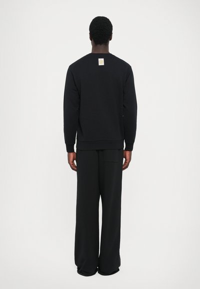 Schwarzer Sweatshirt mit Rundhalsausschnitt, gerippten Bündchen und einem kleinen Logopatch auf der Rückseite, kombiniert mit lockeren schwarzen Hosen. Weicher Stoff und simples Design.