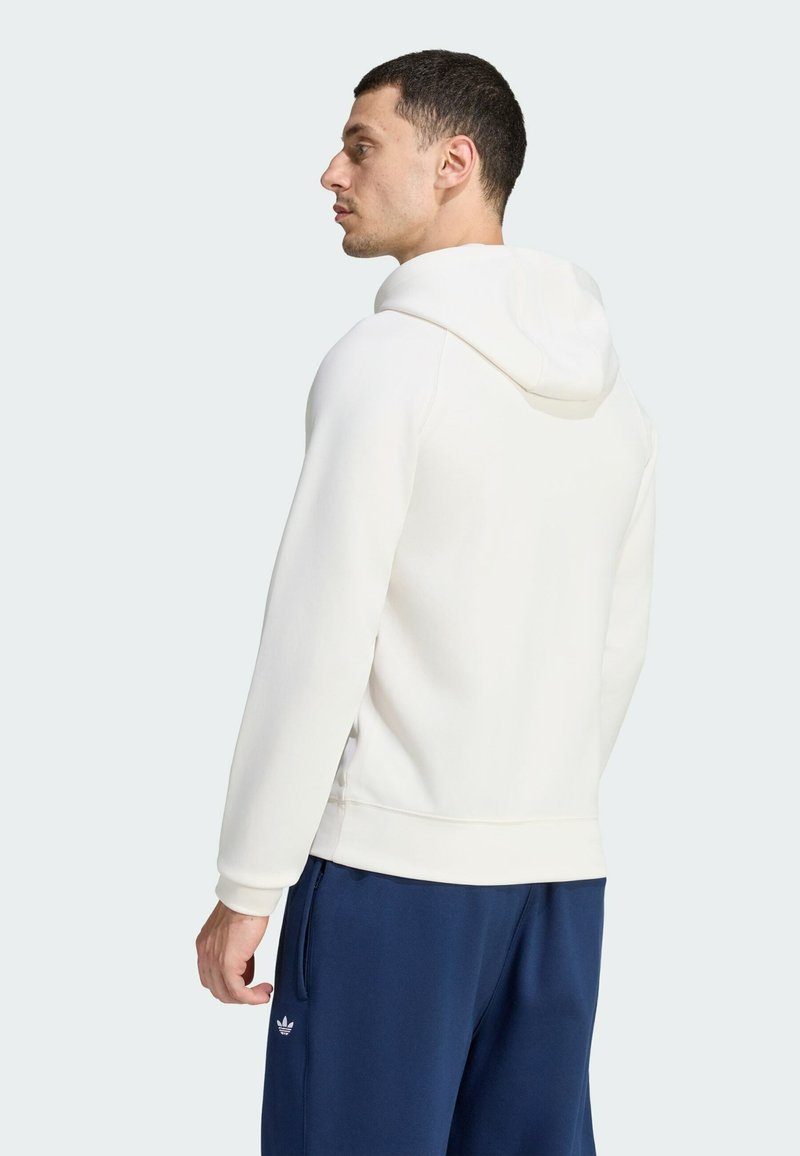 Man facing left wearing a white hoodie and navy blue Adidas pants on a plain light background.