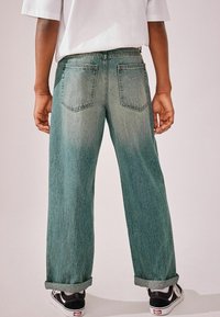 Jean bleu clair à coupe décontractée, avec des ourlets retroussés et deux poches arrière. Porté avec des baskets noir et blanc, texture en denim lisse.
