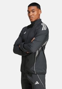 Schwarze Sportjacke aus leichtem Material. Mit weißen Streifen an Schultern und Seiten, einem hohen Kragen und einem Frontreißverschluss.