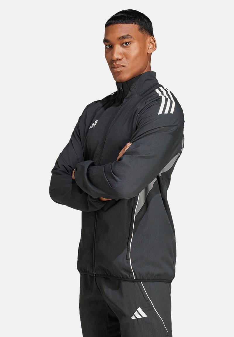 Schwarze Sportjacke aus leichtem Material. Mit weißen Streifen an Schultern und Seiten, einem hohen Kragen und einem Frontreißverschluss.