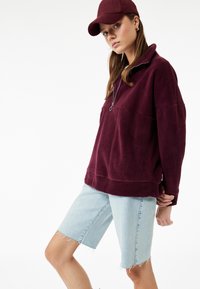 Burgundin värinen fleece-pullovaihtoehto, jossa on neljäsosa vetoketjullinen kaulus ja etutasku, yhdistettynä vaaleansinisiin farkkushortsihousuihin, joissa on risat reunat.