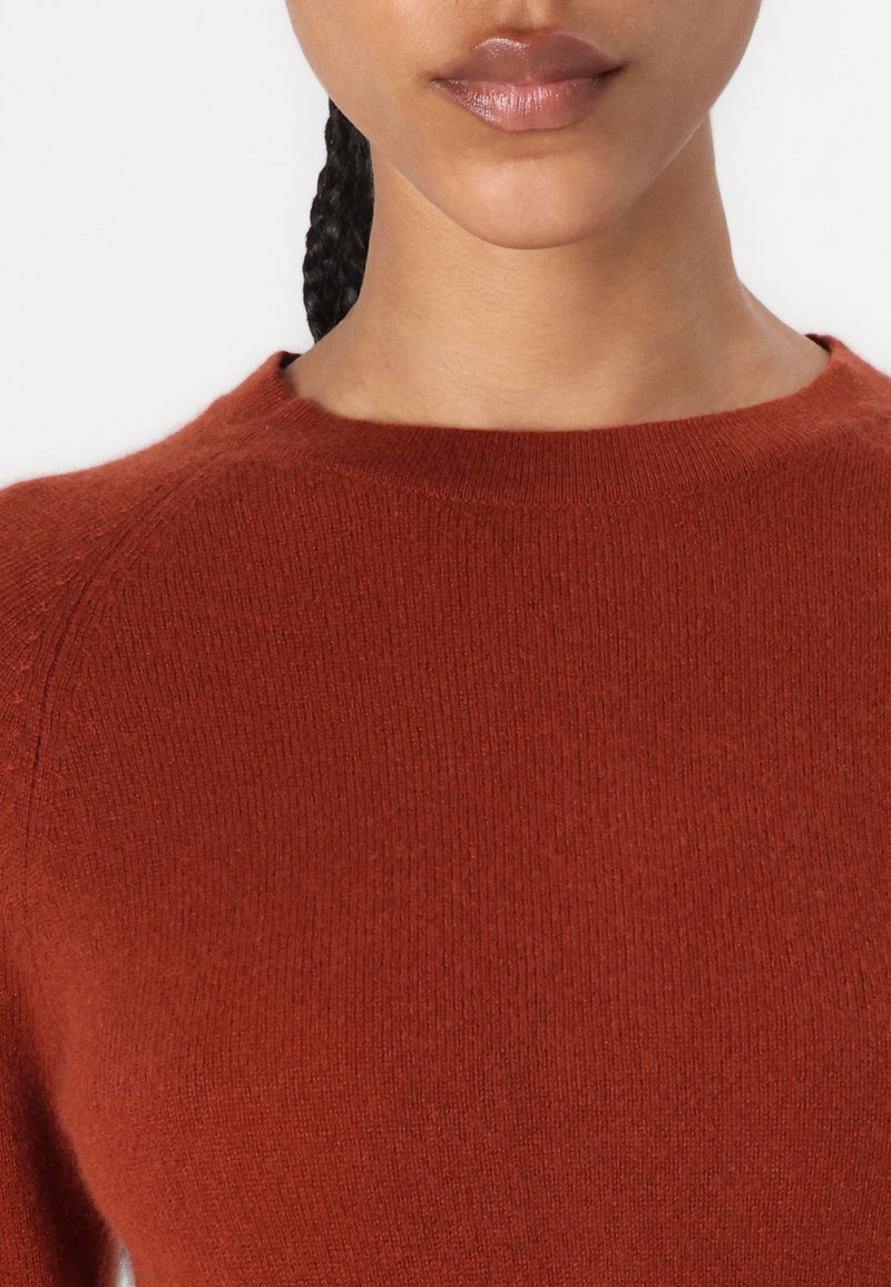 Sweter w kolorze rdzy, dziany, z okrągłym dekoltem i rękawami raglanowymi, charakteryzujący się miękką teksturą i równym ściegiem.