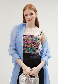 Top corto con textura floral multicolor, abrigo transparente azul claro, pantalones negros de cintura alta y un bolso plateado con diseño recogido.