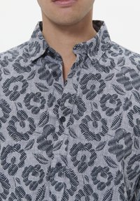 Cămașă cu mânecă scurtă dintr-un material gri deschis, cu un model floral și de frunze în nuanțe de negru, având un guler cu butoane și două butoane închise pe fața cămășii.
