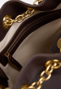 Innenraum einer braunen Lederhandtasche mit goldfarbenen Beschlägen, einem Reißverschlussfach und einem beigen Futter mit eingeprägtem Markennamen "MARC CAIN".