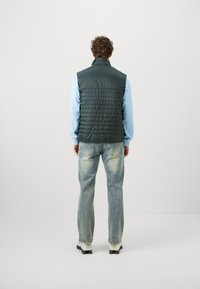 Mørkegrøn dunvest med høj krave, quiltet struktur, båret over en lys blå langærmet skjorte, kombineret med slidte jeans.