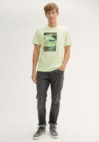 Lichtgroene T-shirt met korte mouwen en een grafische print van geometrische vormen. Gecombineerd met donkergrijze jeans en grijze sneakers.
