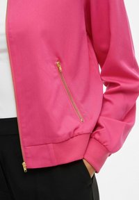 Rosa zip-jacka med gulddragkedjedetalj, bärs över vit topp och svarta byxor, med handen placerad i byxfickan.