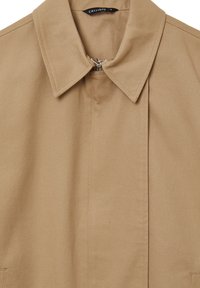 Giacca in cotone color sabbia con colletto a punta, chiusura frontale con zip e design minimalista. Tessuto liscio con sottili dettagli di cucitura.